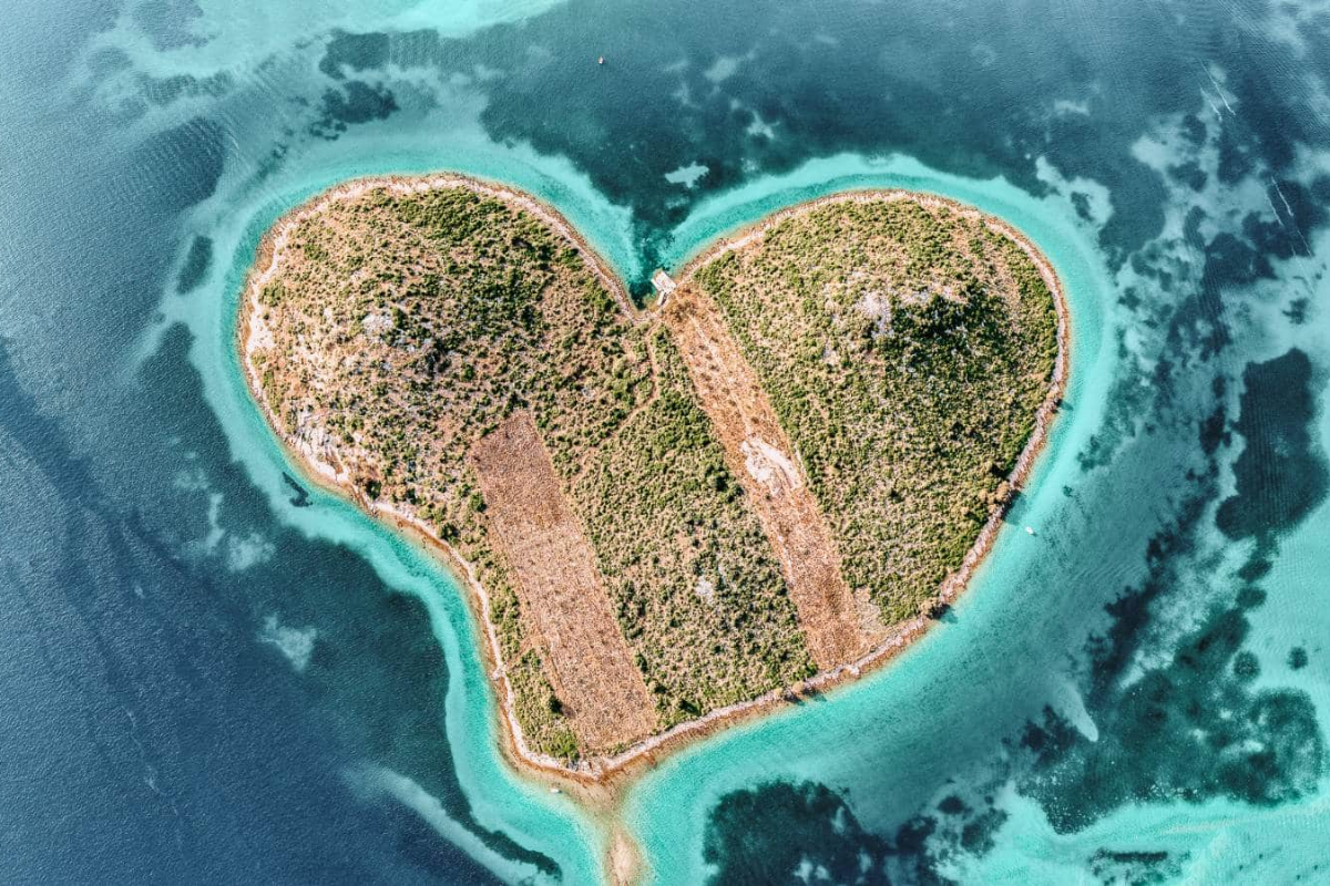 An aerial photograph of a heart-shaped island with green vegetation and sandy beaches surrounded by turquoise and blue ocean waters