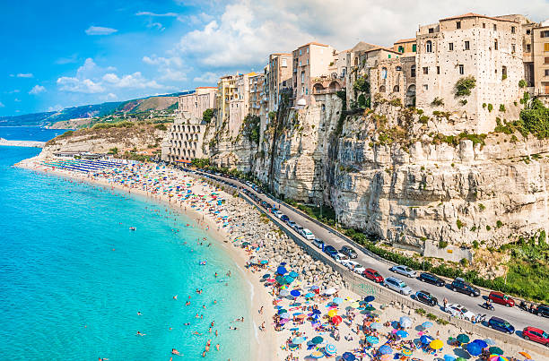 10 Best Places To Visit In Europe In Summer Historic cliffside buildings overlooking a crowded turquoise beach in Tropea, a sun-soaked gem in Italy - Image by iStockphoto.com
